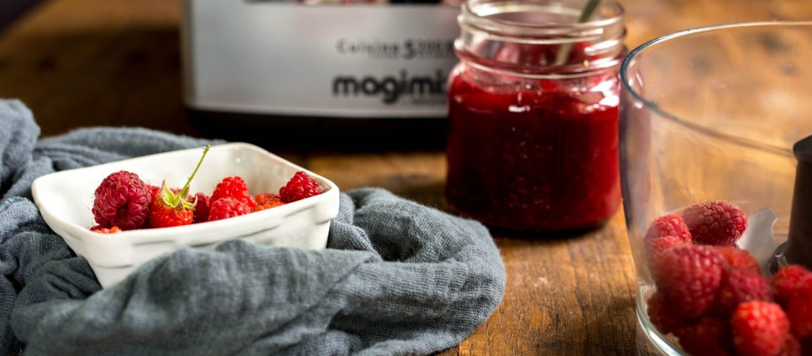 A jar of raspberry jam next to a bowl of raspberries and a Magimix food processor.